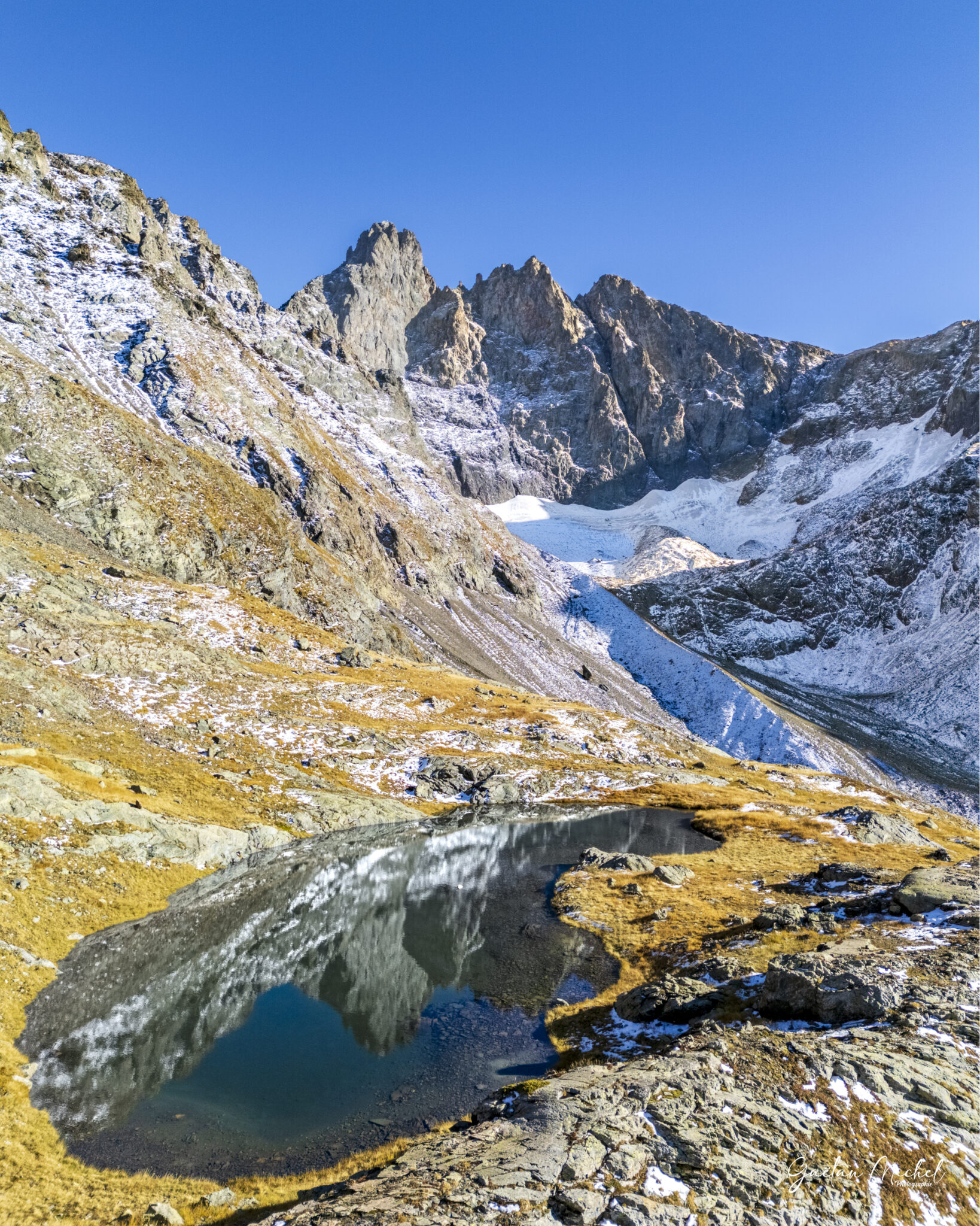 Au cœur du massif de Belledonne, le plus haut sommet se dévoile dans toute sa majesté. Le Grand Pic (2 977 m) se reflète ici dans ce petite lac d’altitude, où l’eau calme capture la pureté du ciel et la rudesse minérale des parois. Les premières neiges d’automne soulignent les reliefs et contrastent avec les teintes dorées des pelouses alpines.
Une scène éphémère, où lumière, roche et glace s’unissent dans une harmonie fragile typique des hauteurs sauvages de Belledonne. Au cœur du massif de Belledonne, le plus haut sommet se dévoile dans toute sa majesté. Le Grand Pic (2 977 m) se reflète ici dans ce petite lac d’altitude, où l’eau calme capture la pureté du ciel et la rudesse minérale des parois. Les premières neiges d’automne soulignent les reliefs et contrastent avec les teintes dorées des pelouses alpines. Une scène éphémère, où lumière, roche et glace s’unissent dans une harmonie fragile typique des hauteurs sauvages de Belledonne.