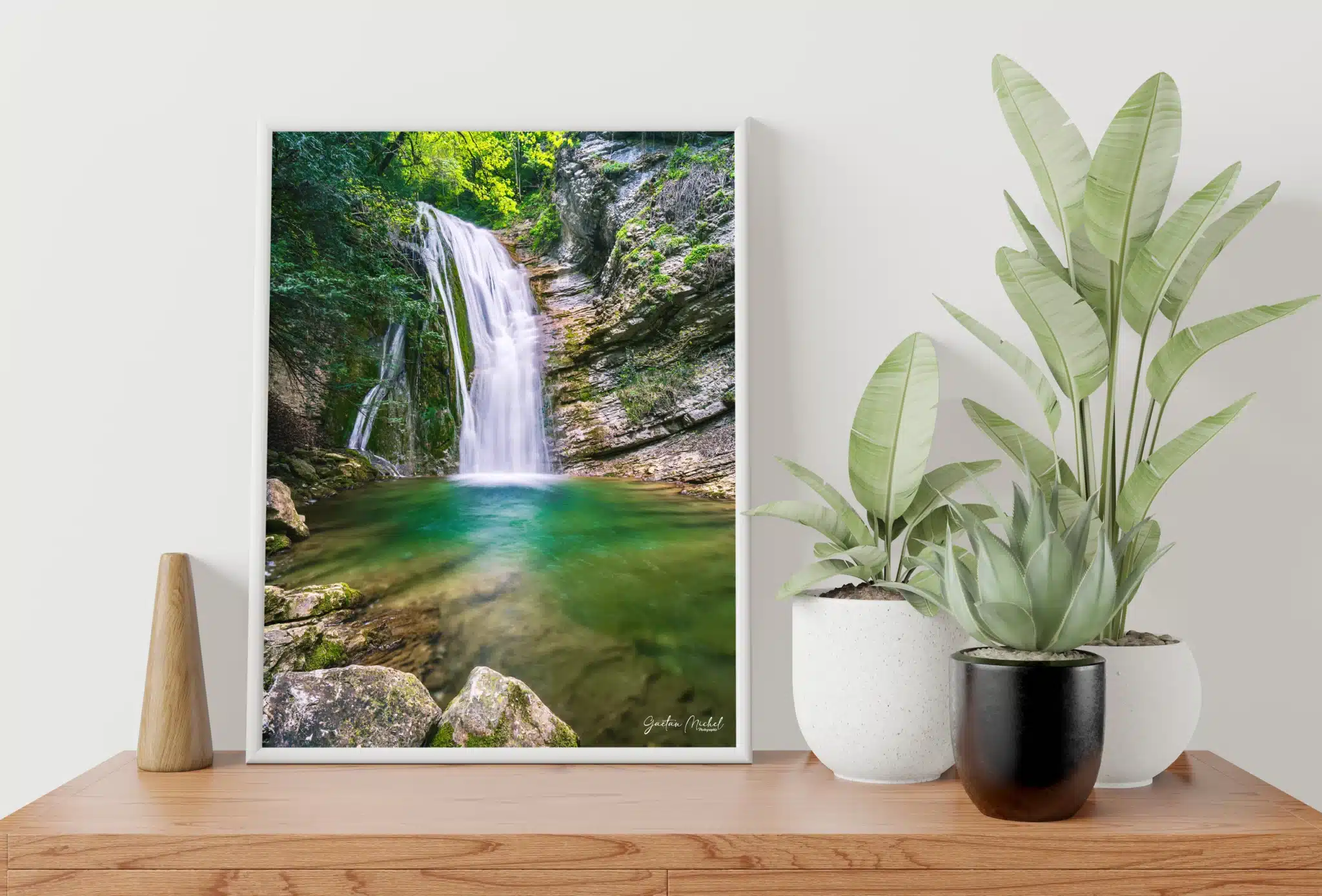 Tirage photo encadré d’une cascade dans le torrent de l’Alloix en Chartreuse, posé sur une étagère en bois clair. L’image montre une cascade avec une eau turquoise et des rochers moussus, entourée de végétation alpine. À côté, deux plantes vertes en pot (un aloe vera et une plante à larges feuilles) ajoutent une touche naturelle à la scène.