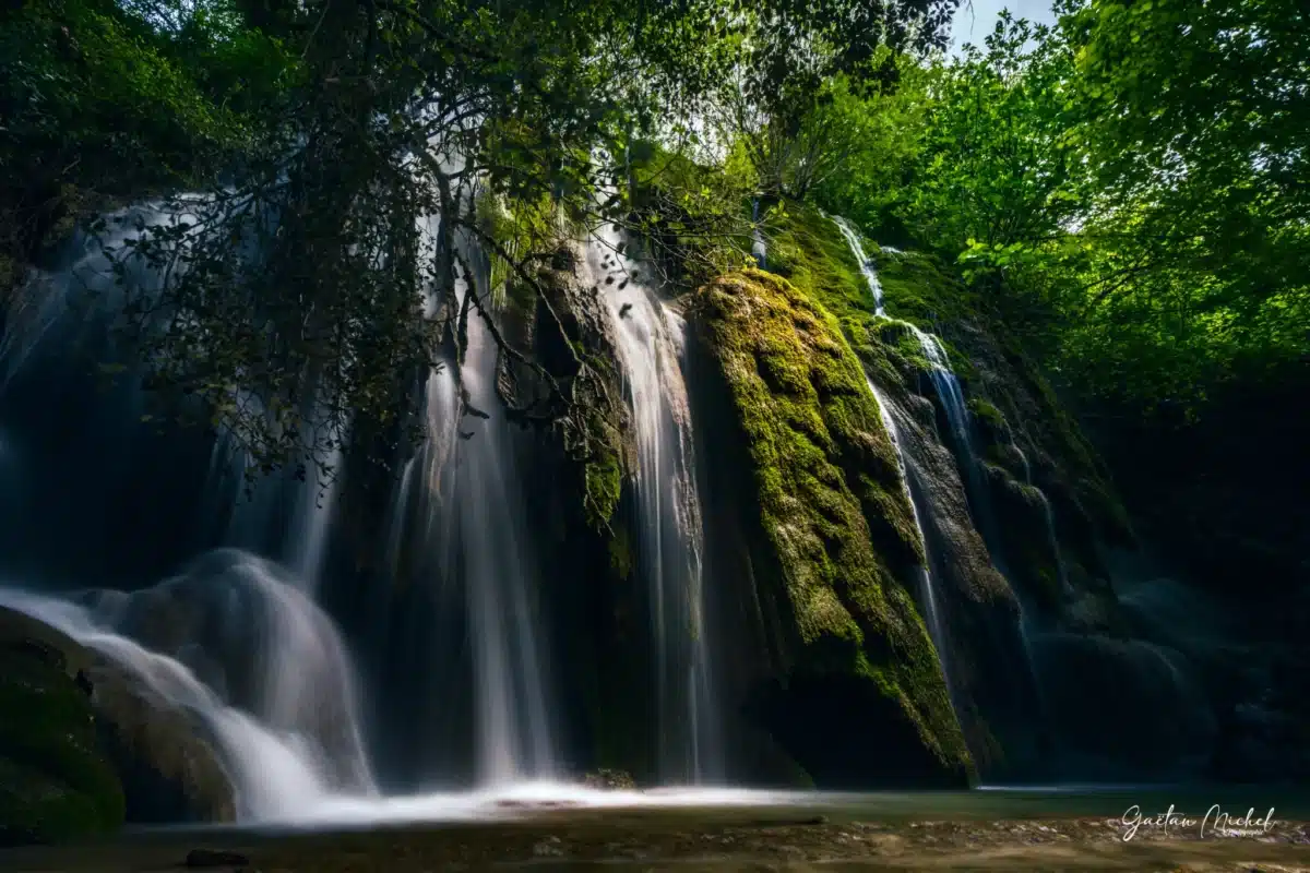 Cascade d’Aubres dans les Baronnies, avec une eau cristalline dévalant des rochers couverts de mousse verte. La lumière naturelle filtre à travers les arbres, créant des jeux d’ombre et de lumière sur l’eau et la végétation dense des Baronnies.