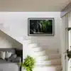 Tirage photo encadré d’une cascade en forêt avec eau cristalline et rochers couverts de mousse verte, accroché au mur d’un escalier blanc. En dessous, un canapé gris avec des coussins à motifs circulaires et une plante verte en pot. Lumière naturelle entrant par une fenêtre à droite.