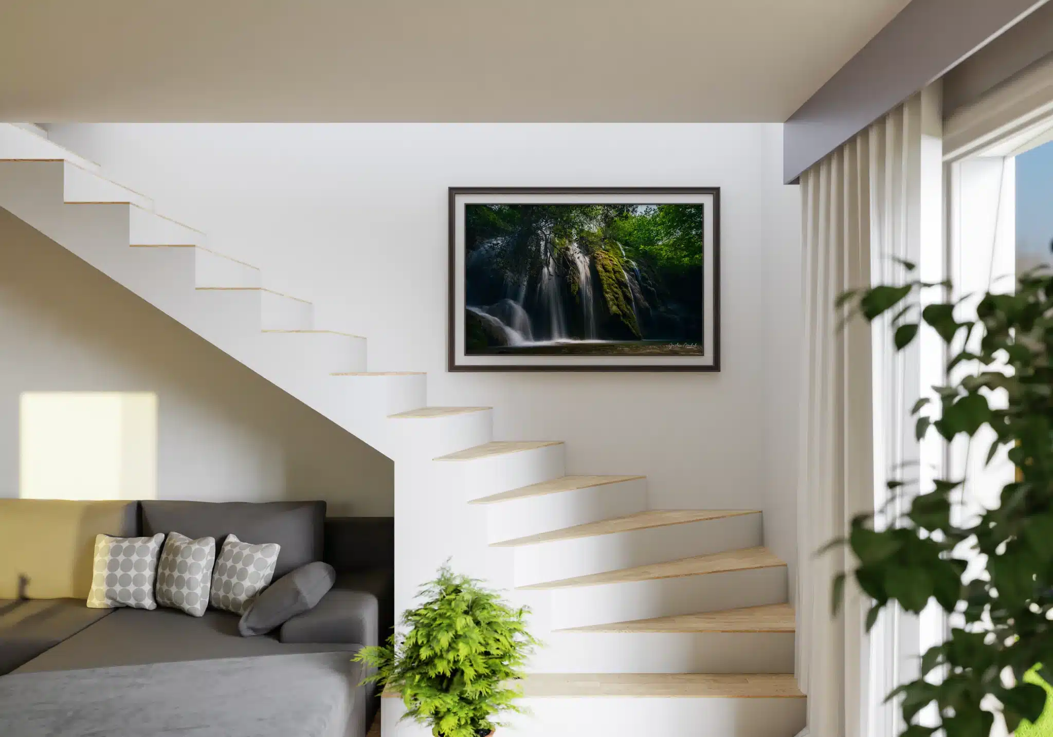 Tirage photo encadré d’une cascade en forêt avec eau cristalline et rochers couverts de mousse verte, accroché au mur d’un escalier blanc. En dessous, un canapé gris avec des coussins à motifs circulaires et une plante verte en pot. Lumière naturelle entrant par une fenêtre à droite.