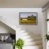 Tirage photo encadré d’un paysage alpin avec lac et montagnes, accroché au mur d’un escalier blanc. En dessous, un canapé gris avec des coussins à motifs et une plante verte en pot. Lumière naturelle entrant par une fenêtre à droite.