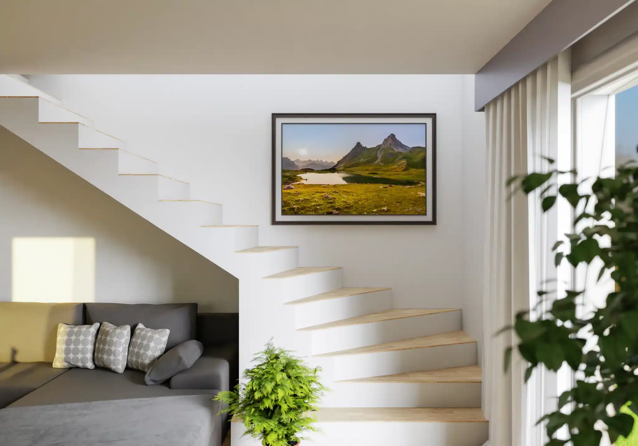Tirage photo encadré d’un paysage alpin avec lac et montagnes, accroché au mur d’un escalier blanc. En dessous, un canapé gris avec des coussins à motifs et une plante verte en pot. Lumière naturelle entrant par une fenêtre à droite.