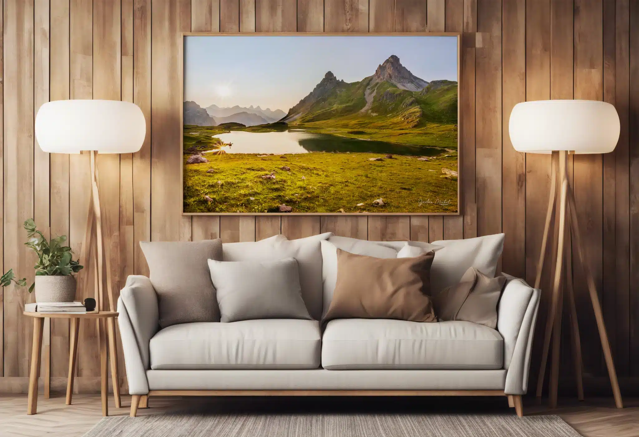 Tirage photo encadré d’un paysage alpin avec lac et montagnes, accroché au mur en bois clair d’un salon. En dessous, un canapé beige avec des coussins, deux lampes sur pied de chaque côté, et une petite table avec une plante verte.