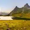 Lac des Cerces à Valloire avec les Aiguilles d’Arves en arrière-plan, reflet doré du soleil sur l’eau, prairies verdoyantes et lumière chaude du coucher ou lever de soleil.