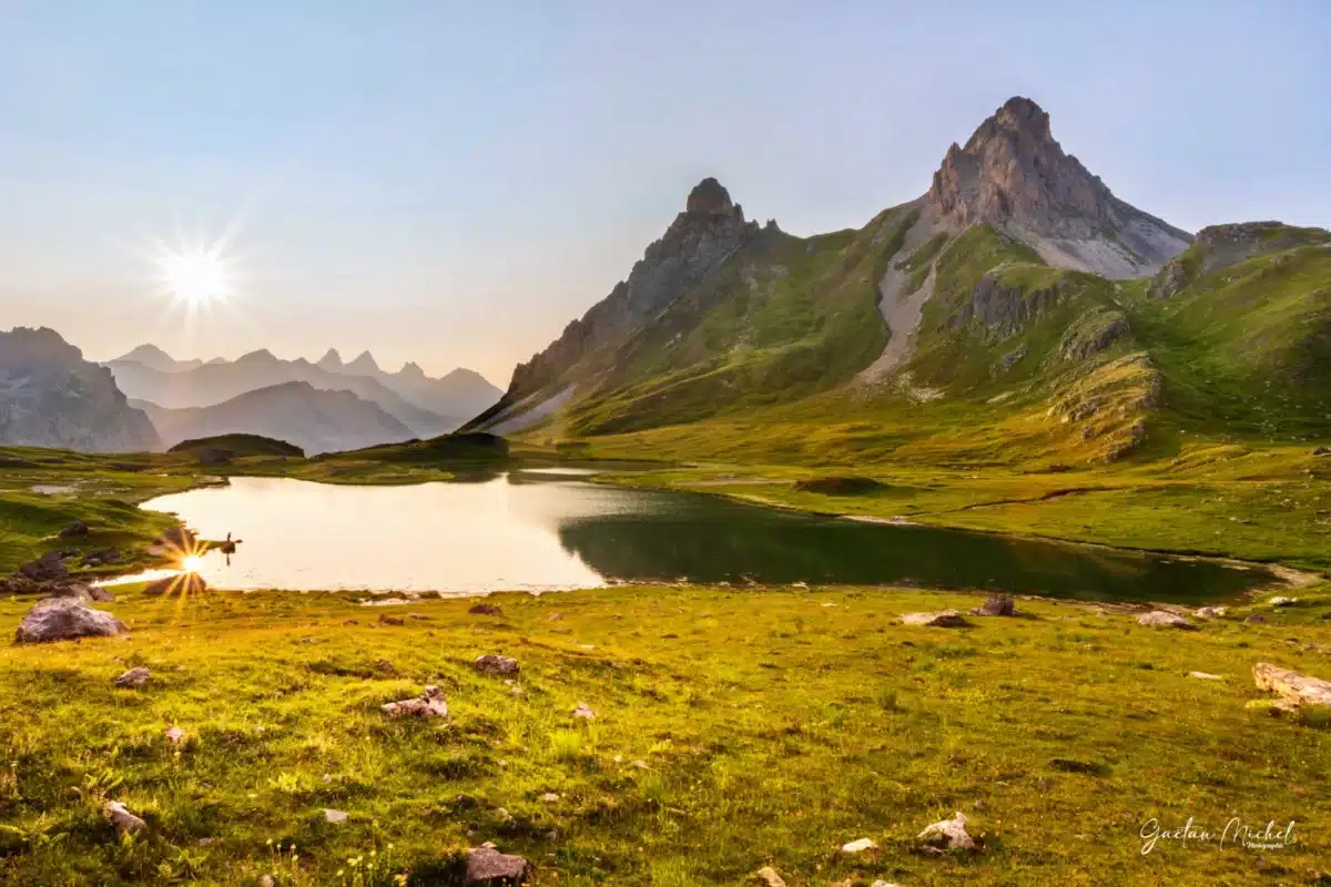 Lac des Cerces à Valloire avec les Aiguilles d’Arves en arrière-plan, reflet doré du soleil sur l’eau, prairies verdoyantes et lumière chaude du coucher ou lever de soleil.